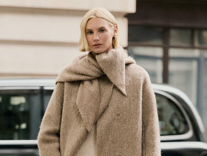 a woman in a beige coat and scarf standing on the street
