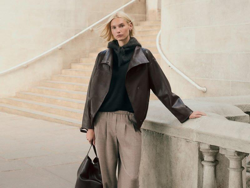 a woman in a black sweater and brown jacket leaning against a wall