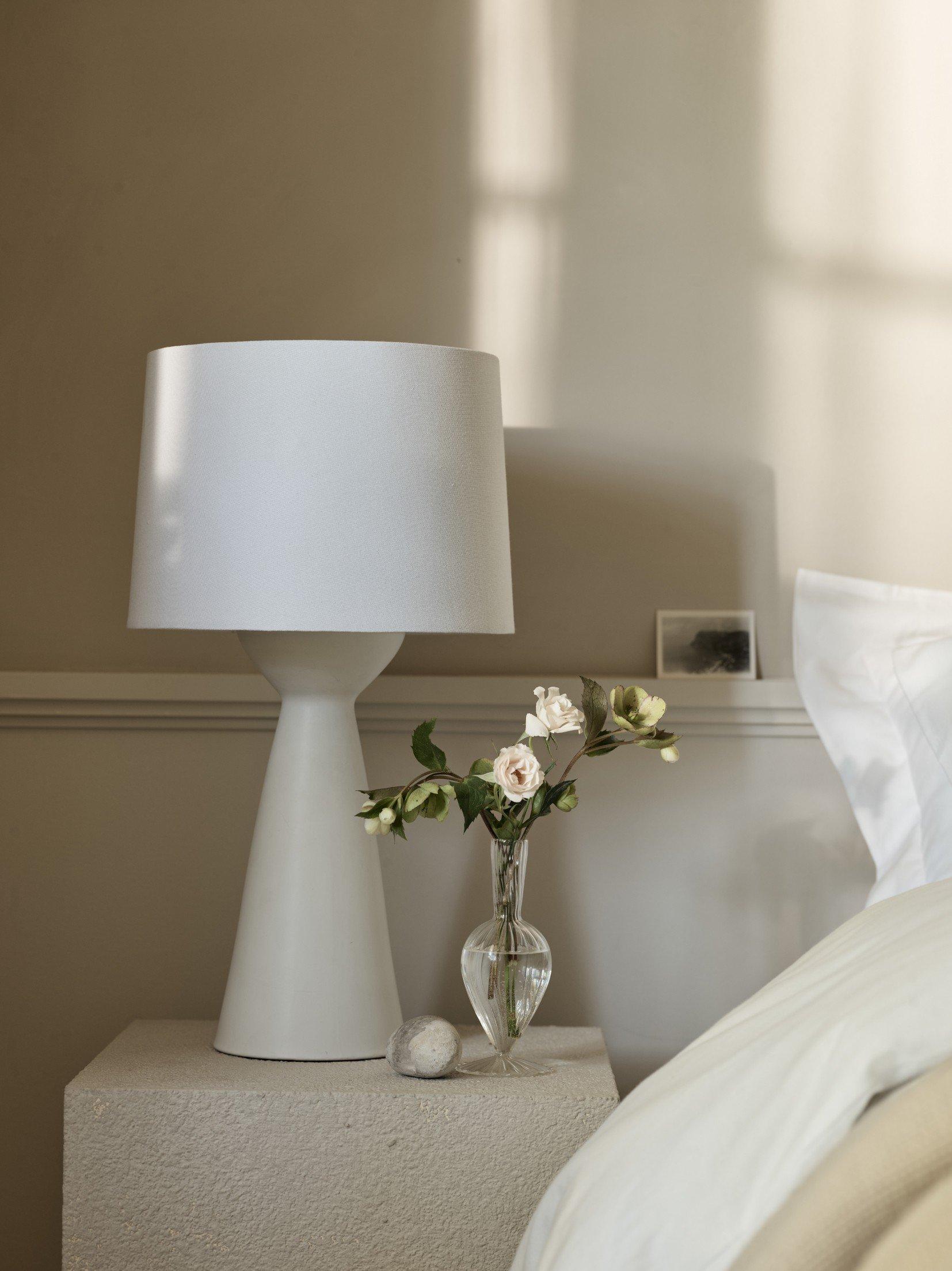 a white bed with a white lamp and a vase of flowers