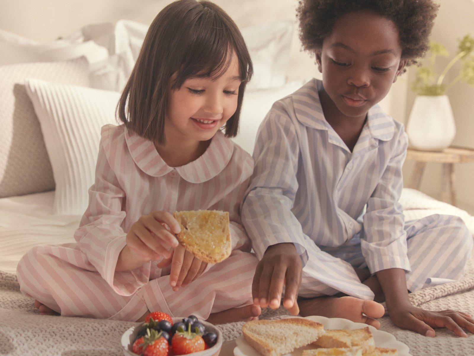 two children in pajamas eating food on a bed