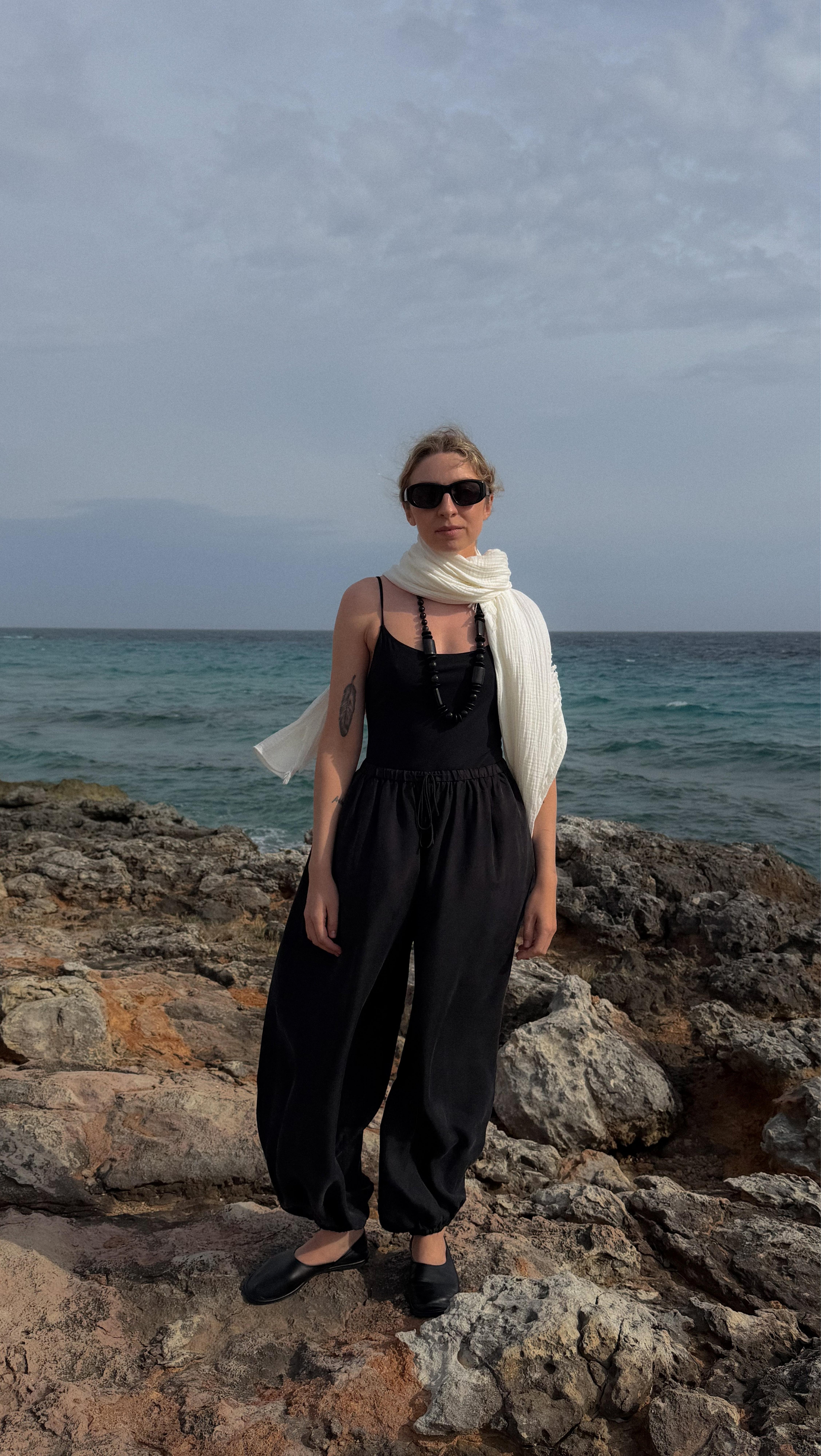 a woman standing on a rocky beach wearing a scarf and sunglasses
