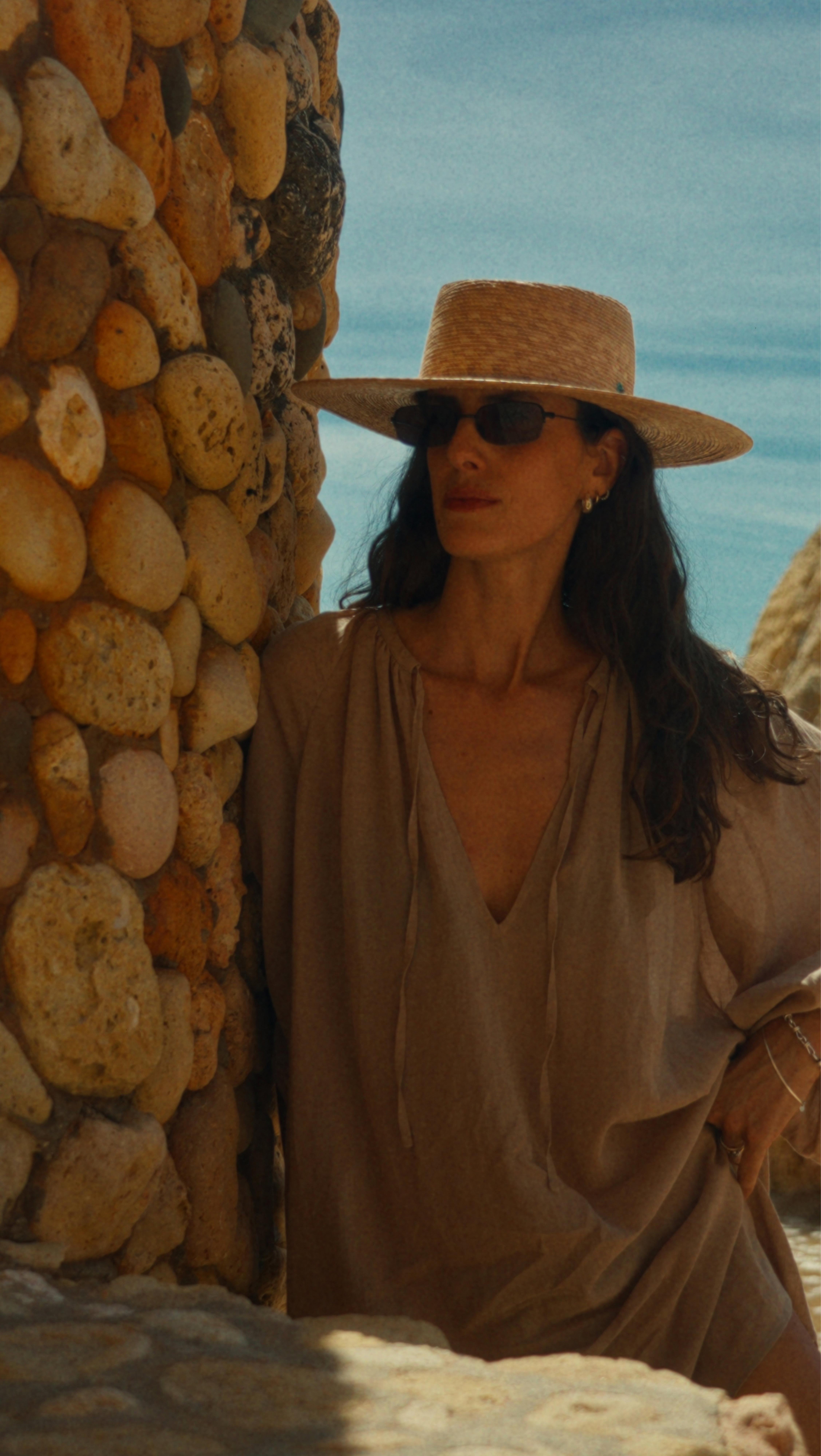 a woman in a hat and sunglasses leaning against a wall