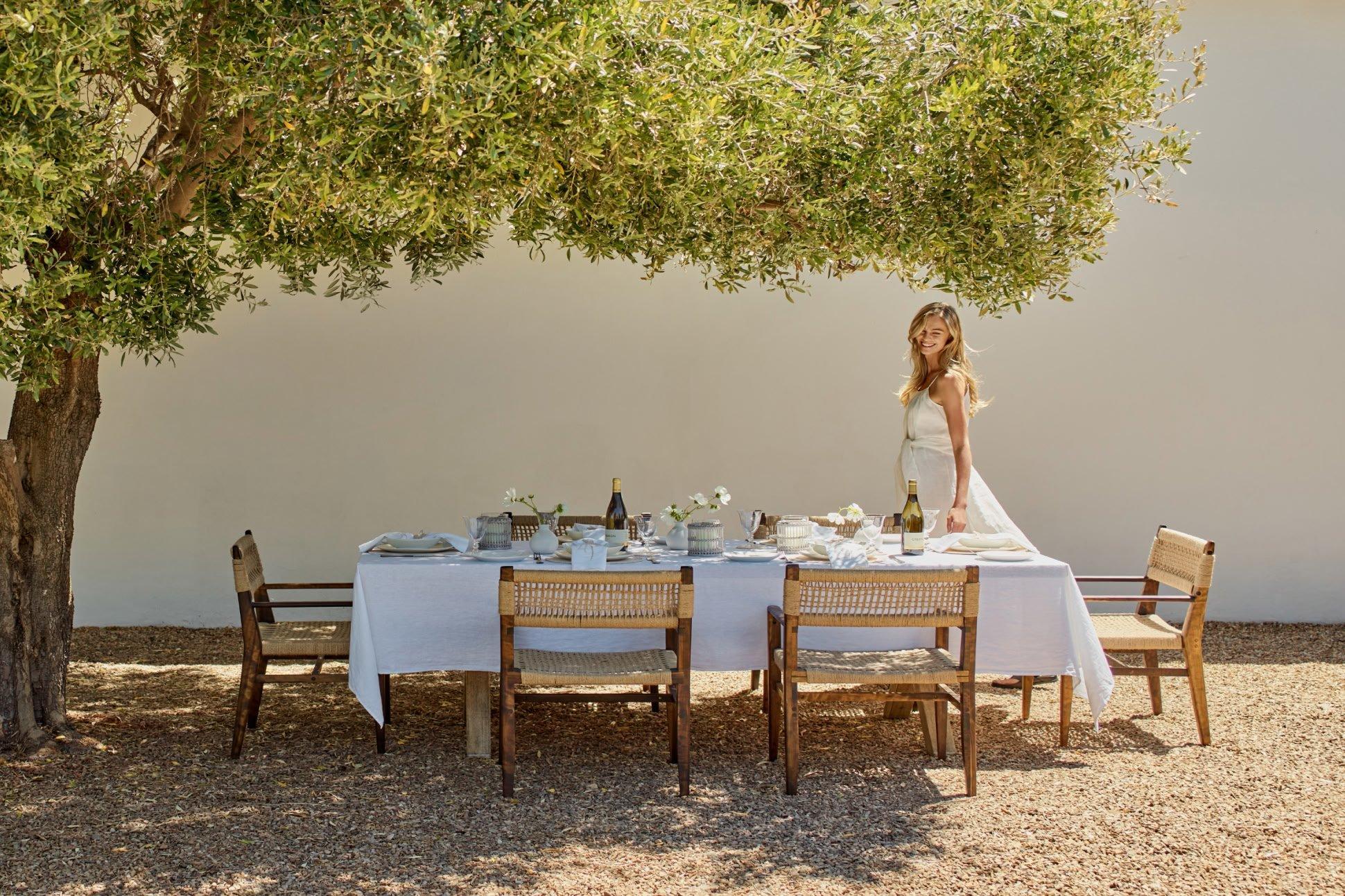 a woman in a white dress standing under a tree with a table set for a dinner party