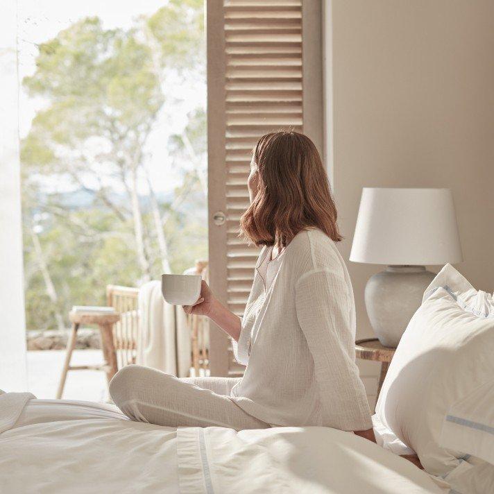 a woman in white sitting on a bed holding a cup