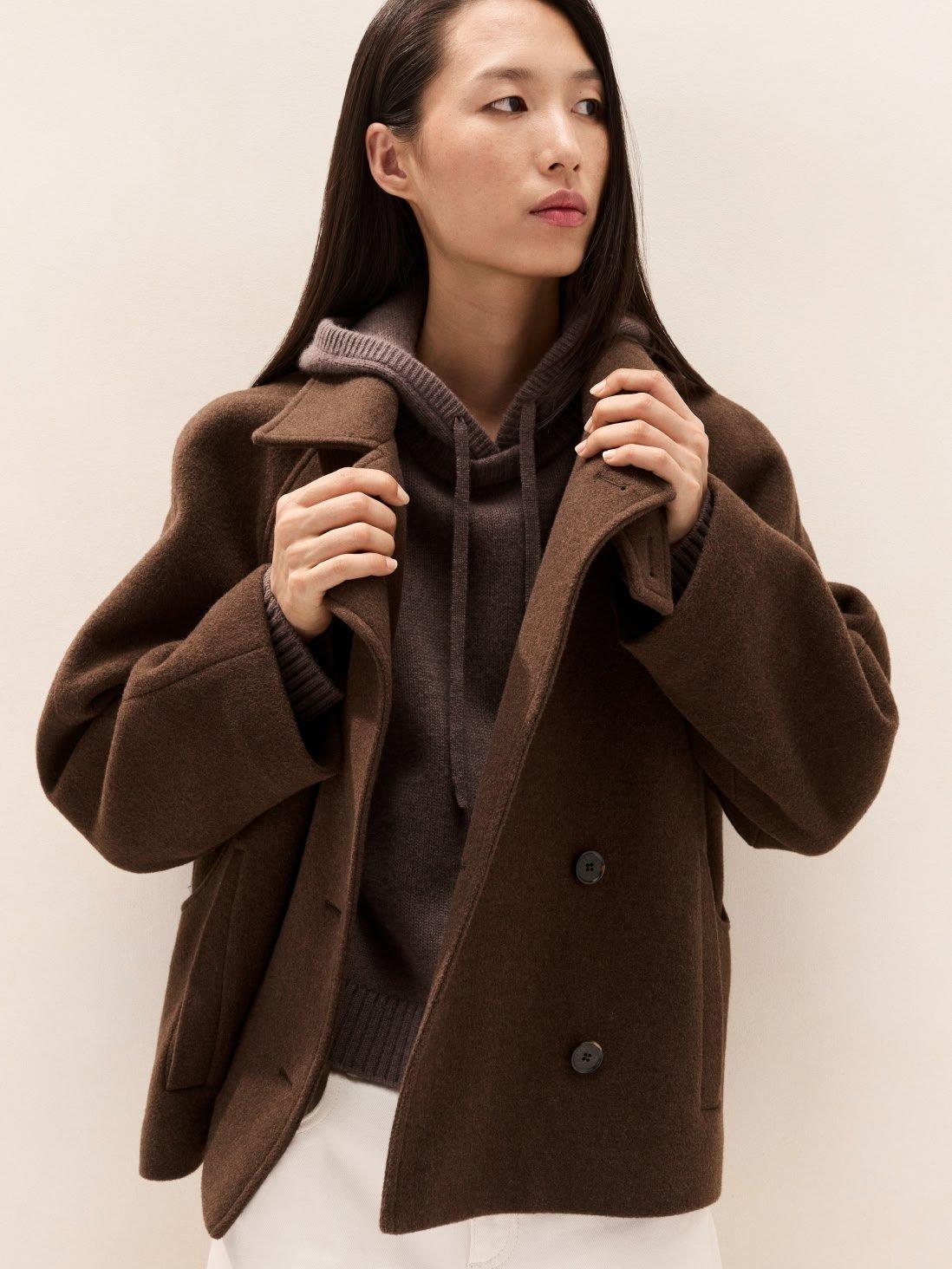 a woman in a brown coat standing against a white wall