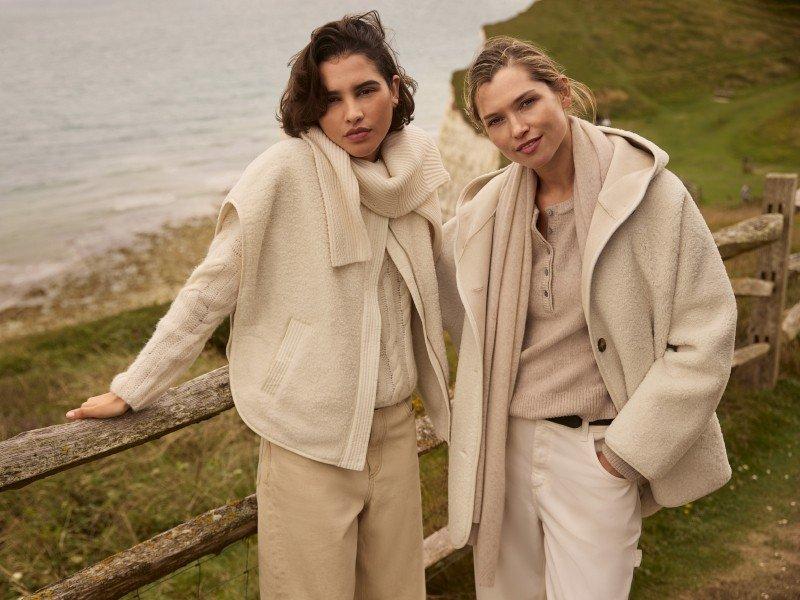 two women in beige coats standing next to a fence by the ocean