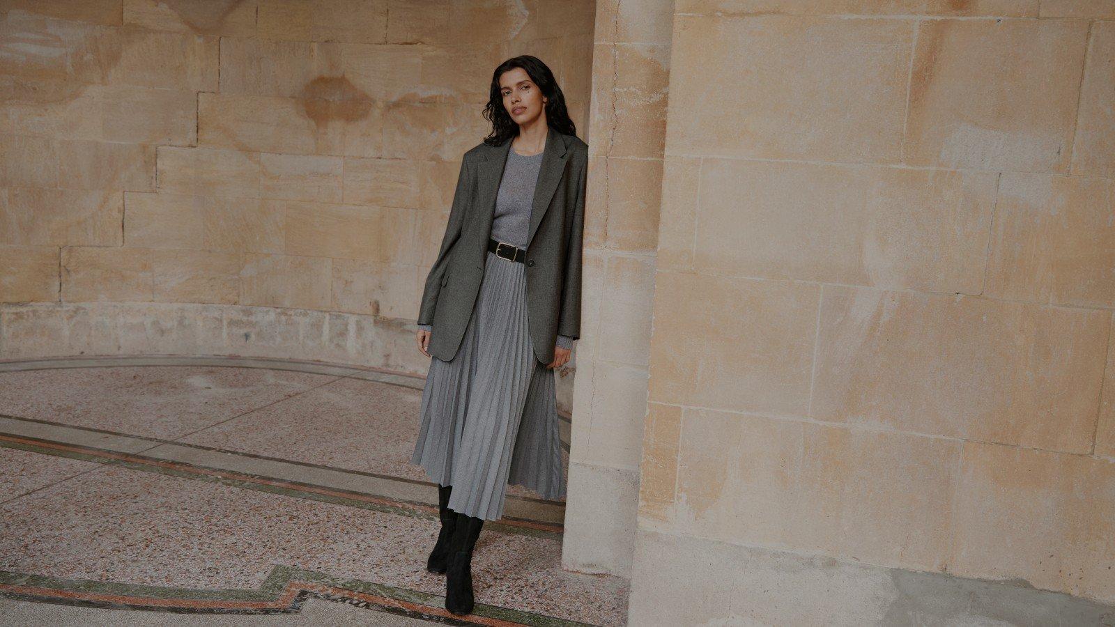 a woman in grey skirt and jumper leaning against a building