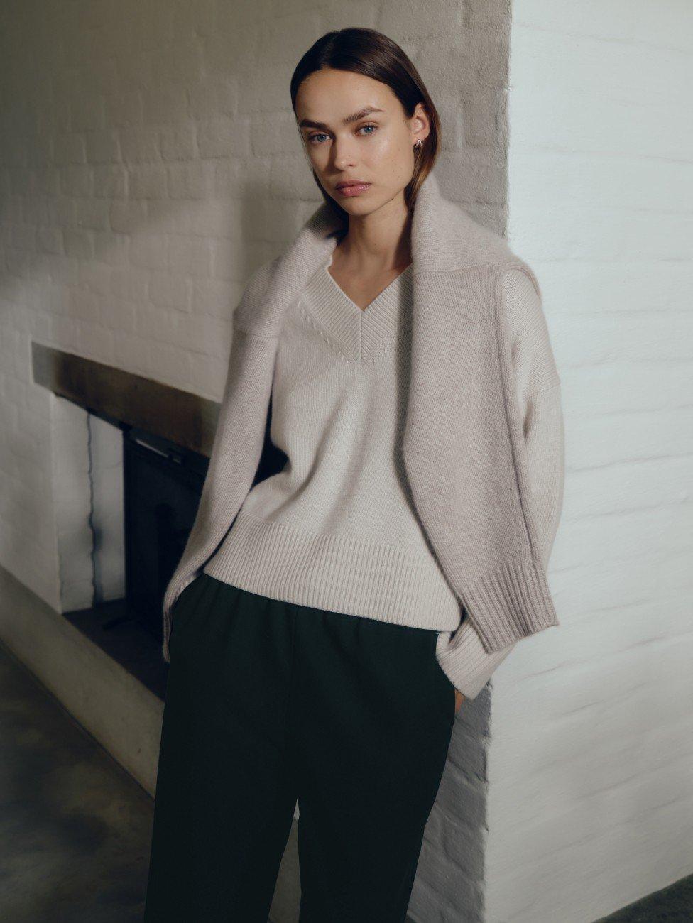 a woman in a sweater and pants standing by a fireplace