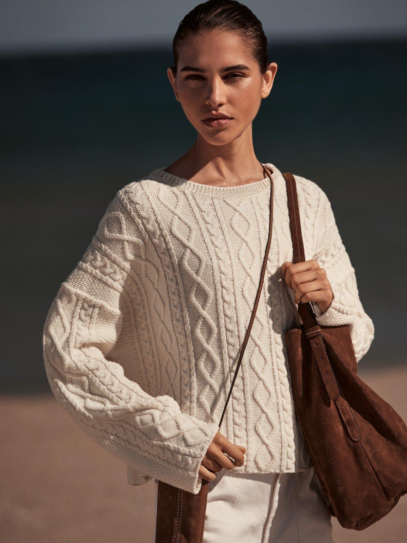 a woman in a white sweater and brown bag on the beach