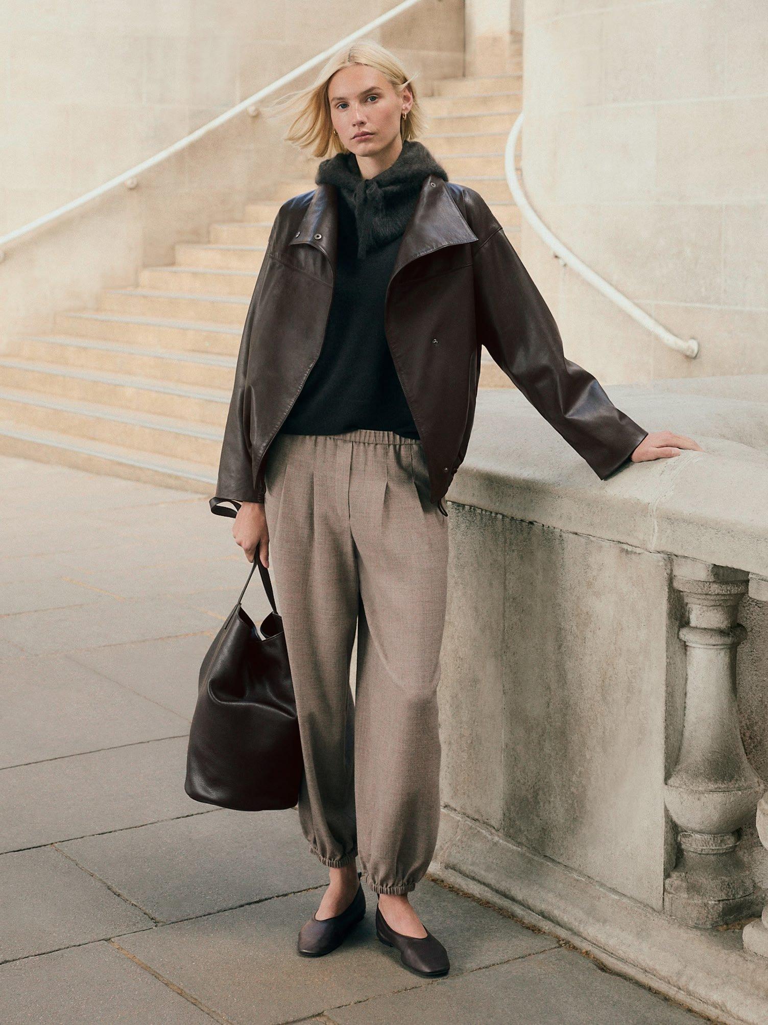 a woman in a jacket and pants leaning against a wall