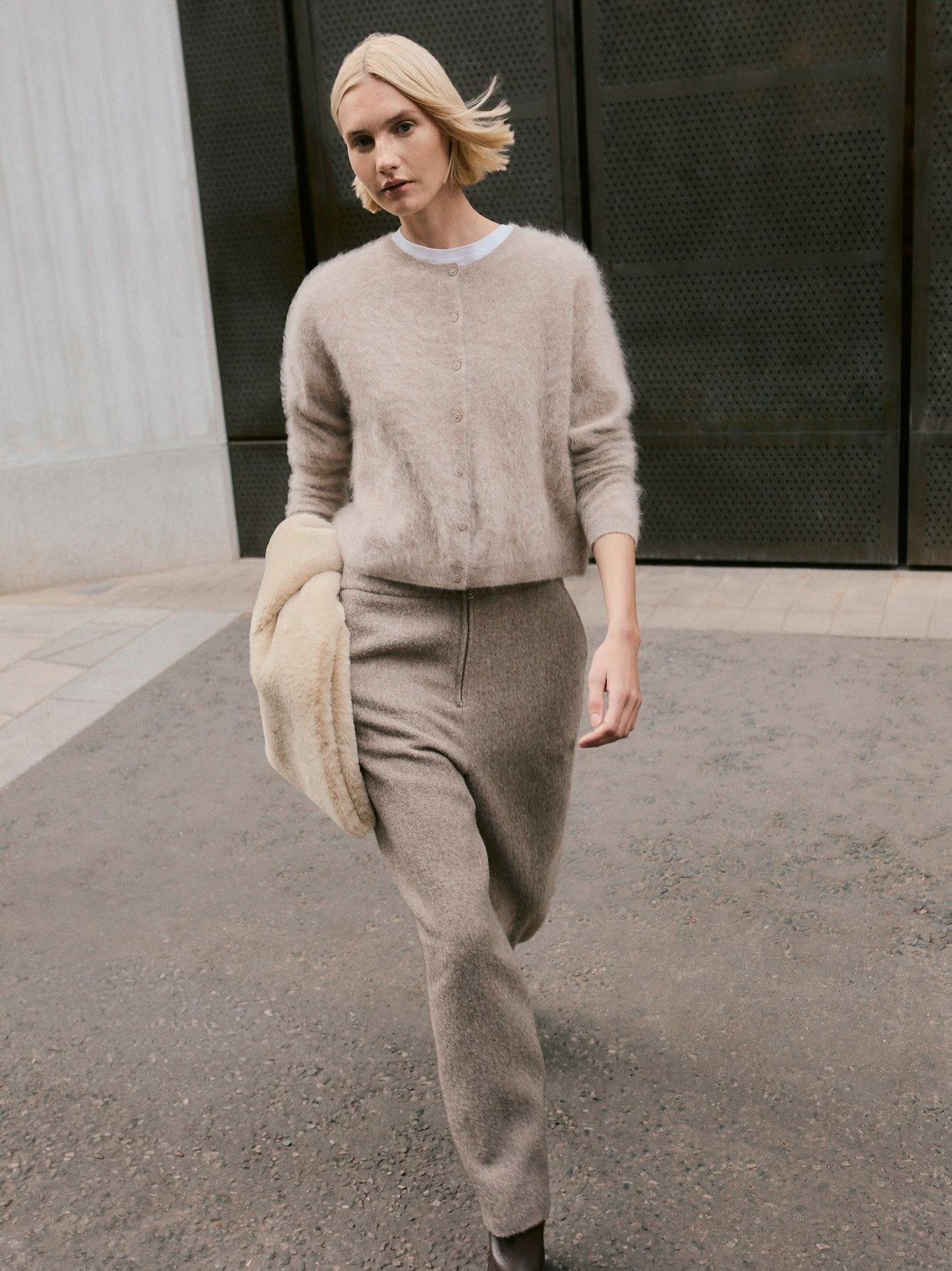 a woman walking down a street in a beige sweater and pants