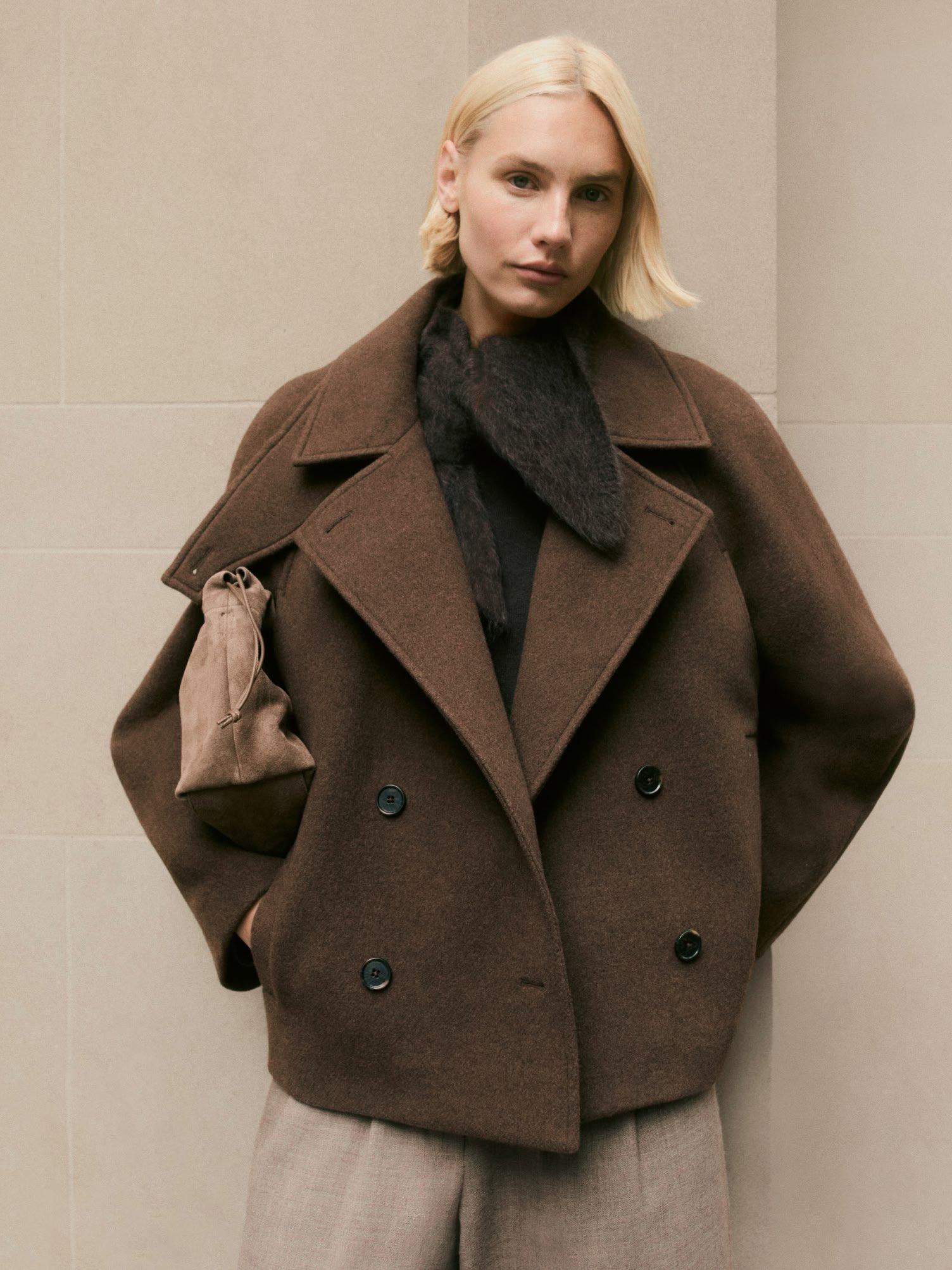a woman in a brown coat leaning against a wall