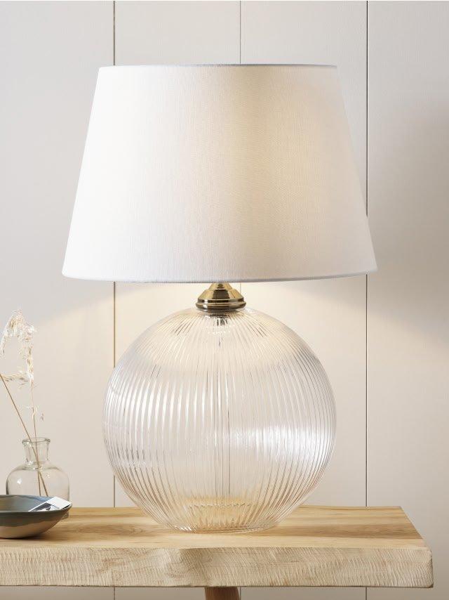 a clear glass table lamp with a white shade on top of a wooden table