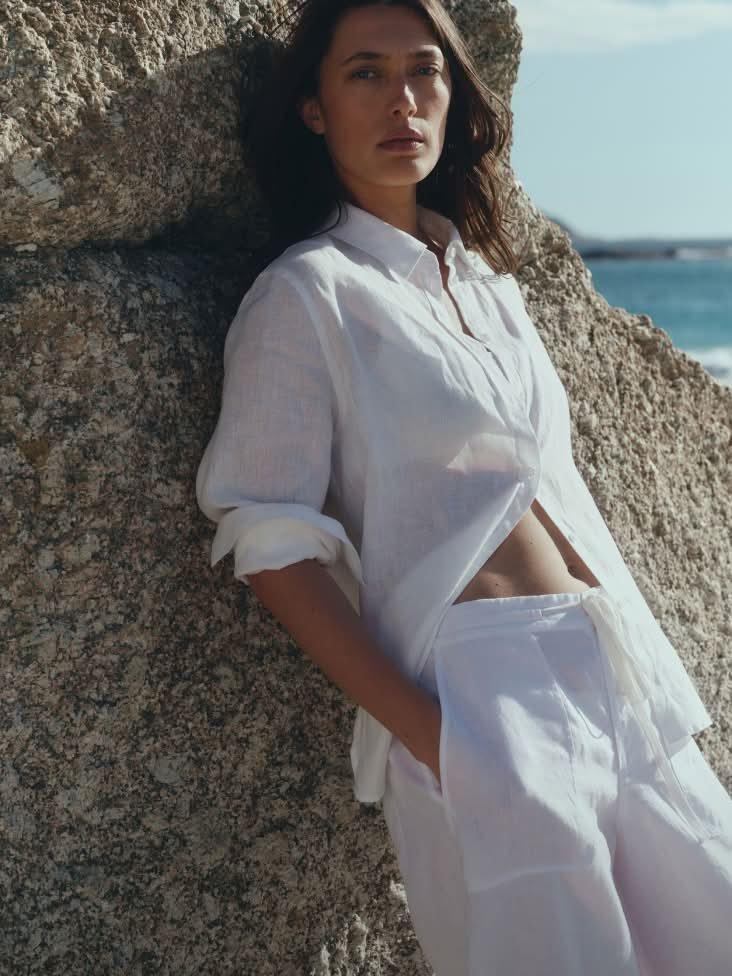 a woman in white pants leaning against a rock by the ocean