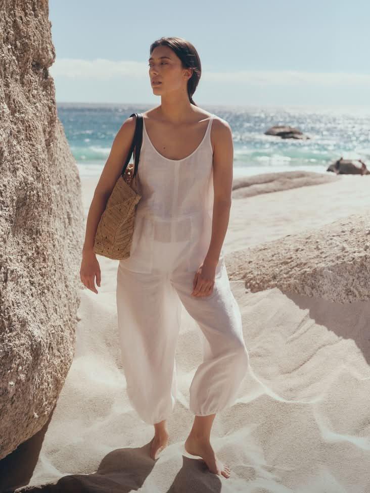 a woman in white jumpsuit standing on beach near large rock