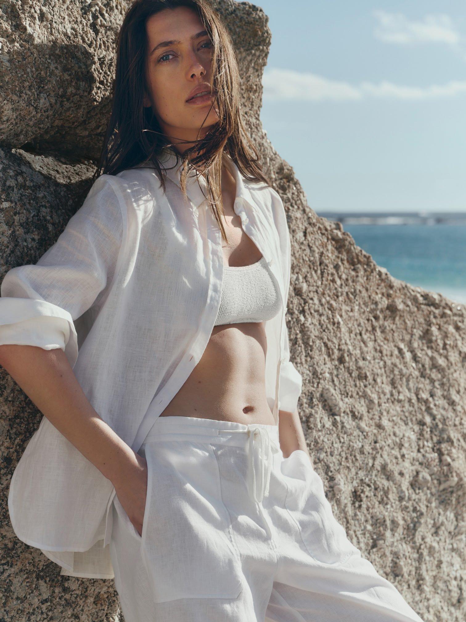 a woman in white pants leaning against a rock by the ocean
