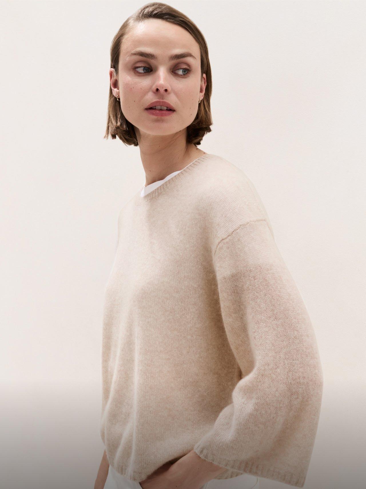 a woman leaning against a wall wearing a beige sweater