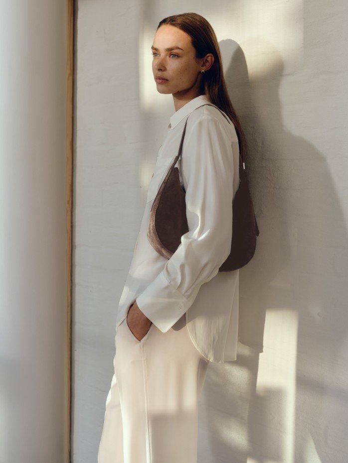 a woman standing against a wall wearing white pants and a brown bag