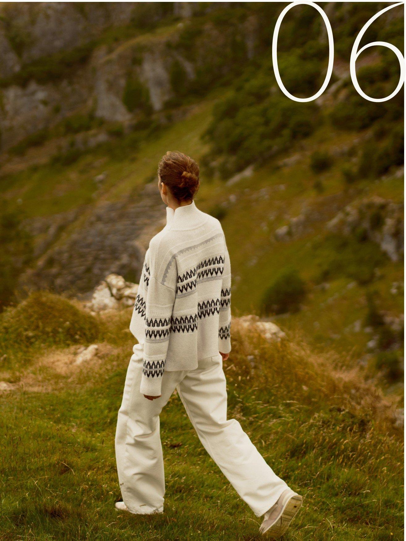 a woman in white pants and jacket walking on grassy hill