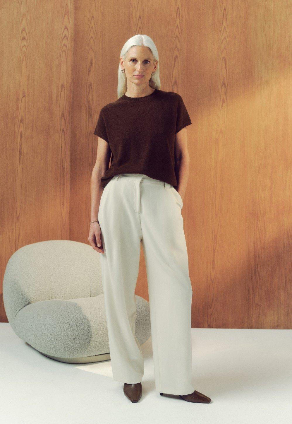 a woman standing in front of a chair wearing a brown shirt and white pants