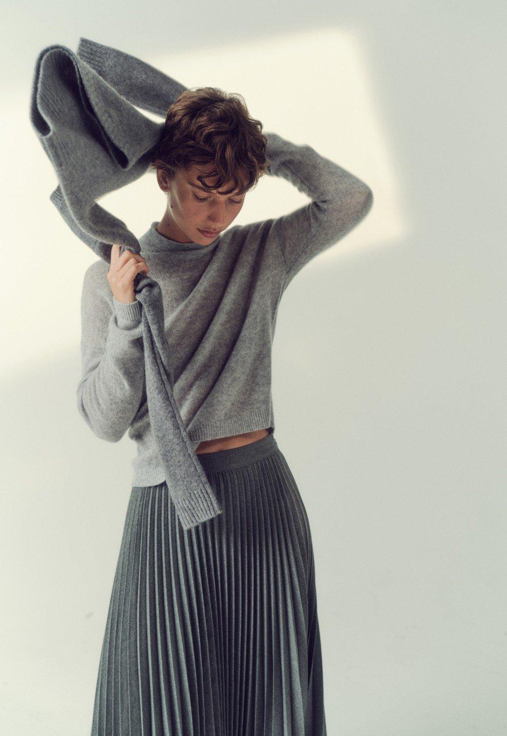 a woman in a grey sweater and a pleated skirt holding a sweater