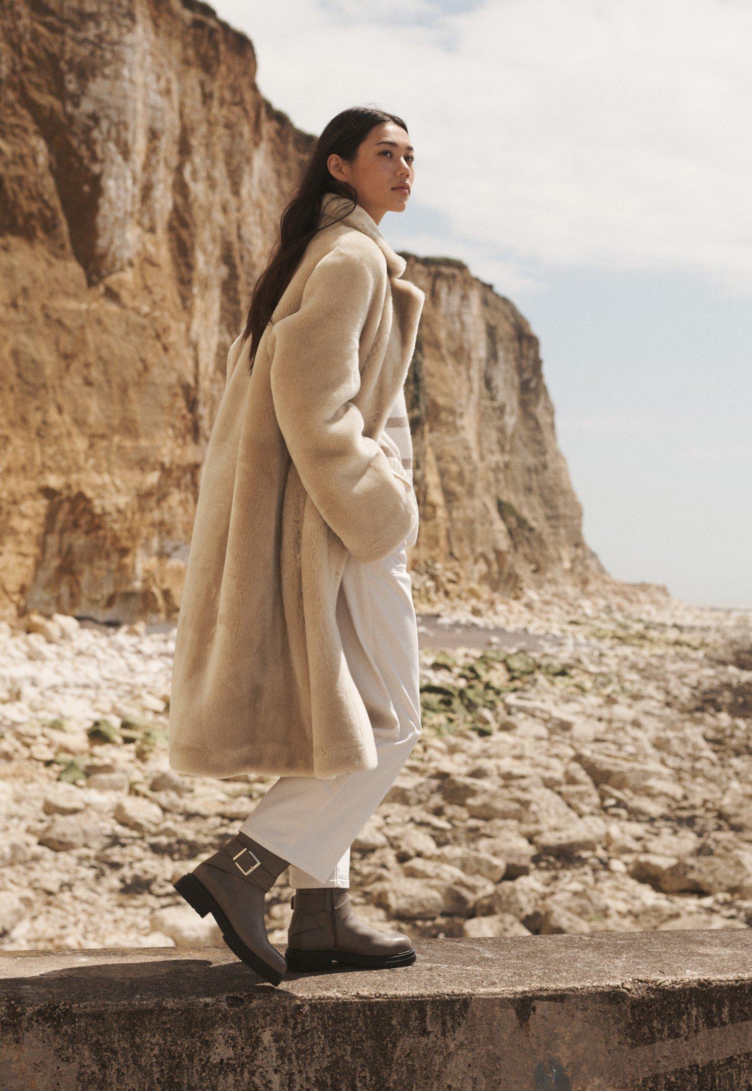 a woman in a fur coat standing on a ledge by the ocean