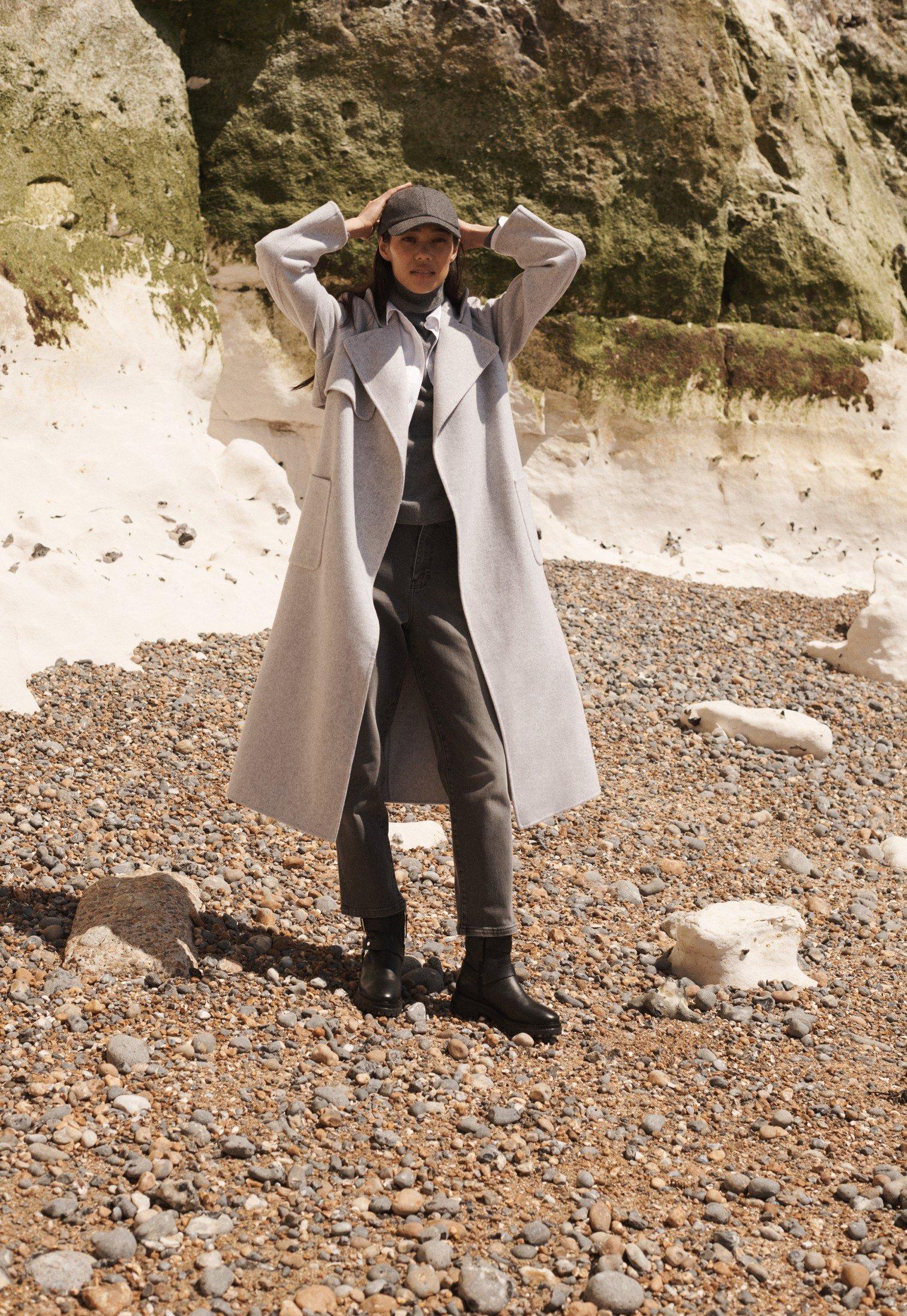 a woman in a long coat standing on a rocky beach