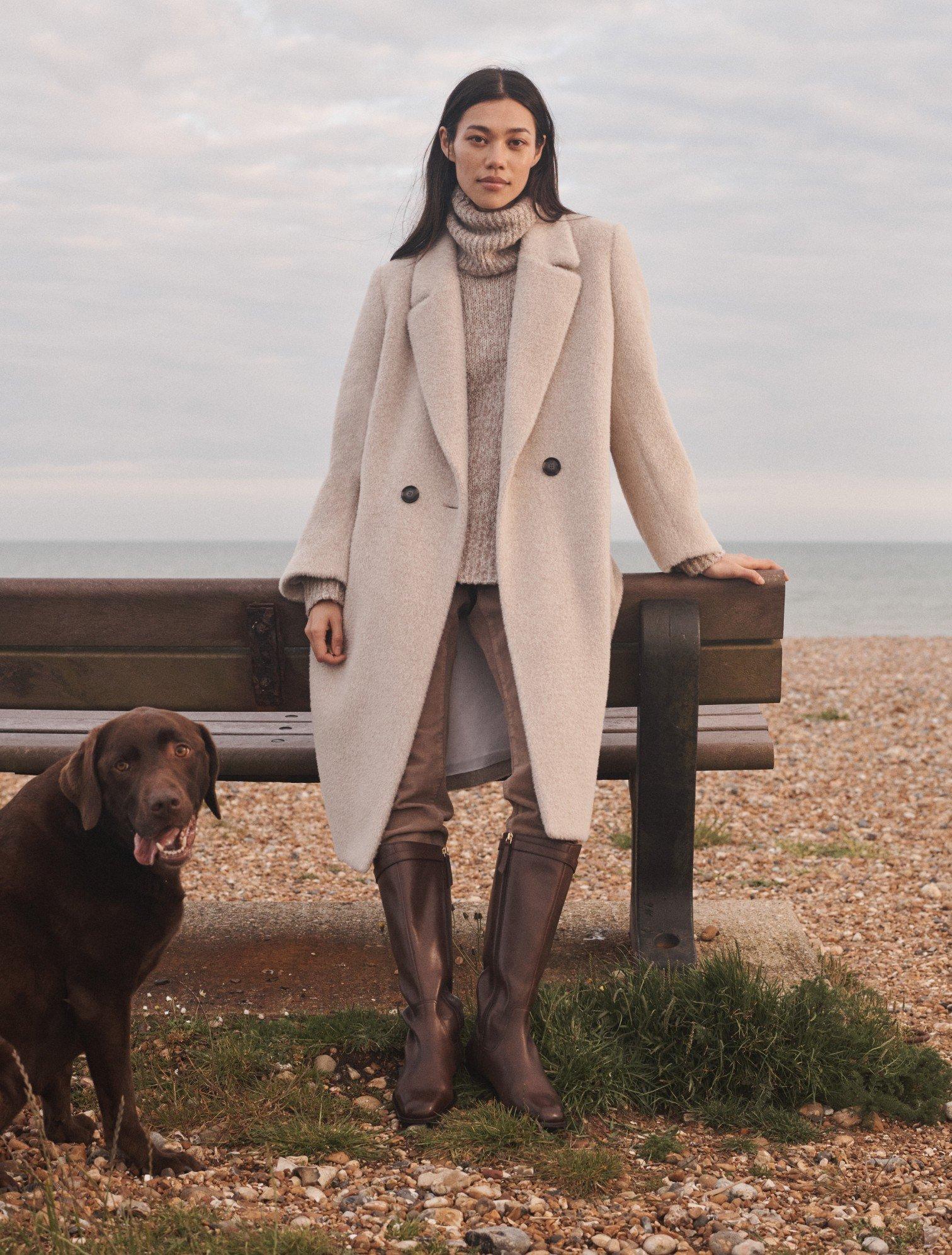 a woman in a coat standing next to a dog on a beach