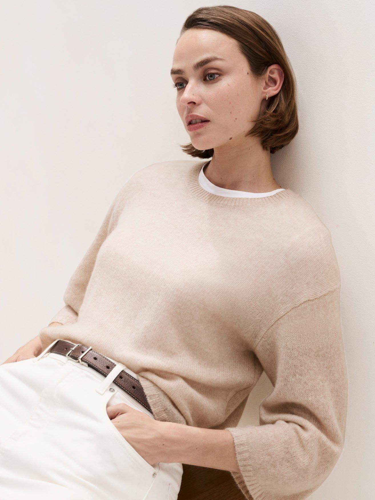 a woman in a beige sweater leaning against a wall