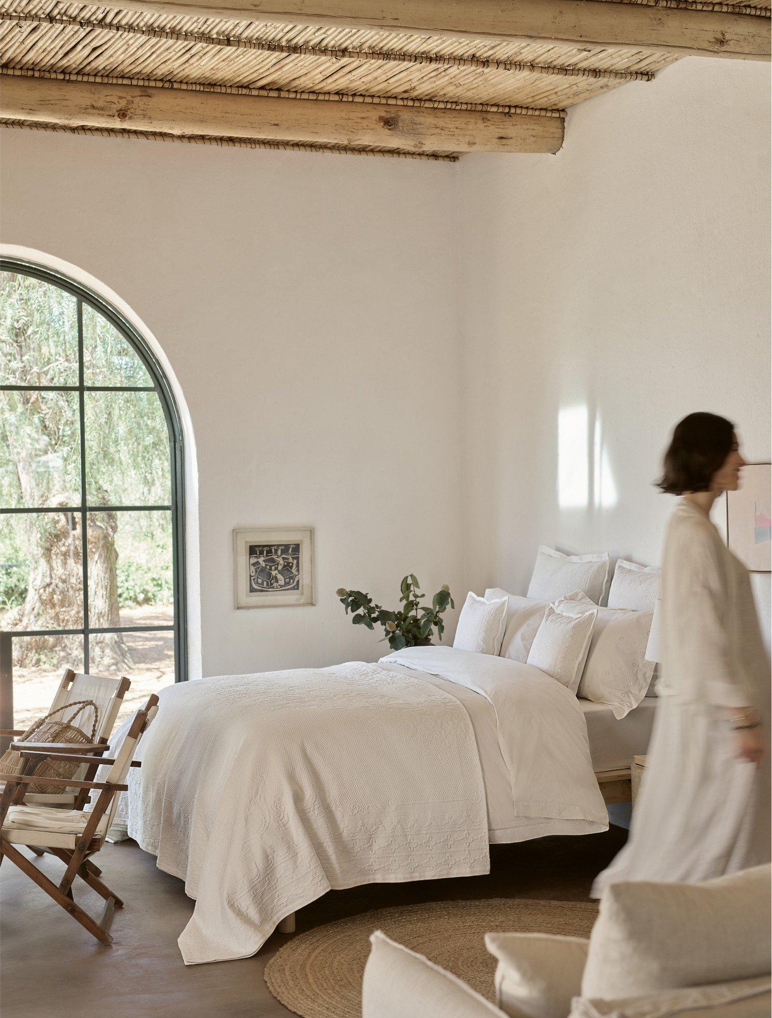 a woman walking through a room with a bed and a window