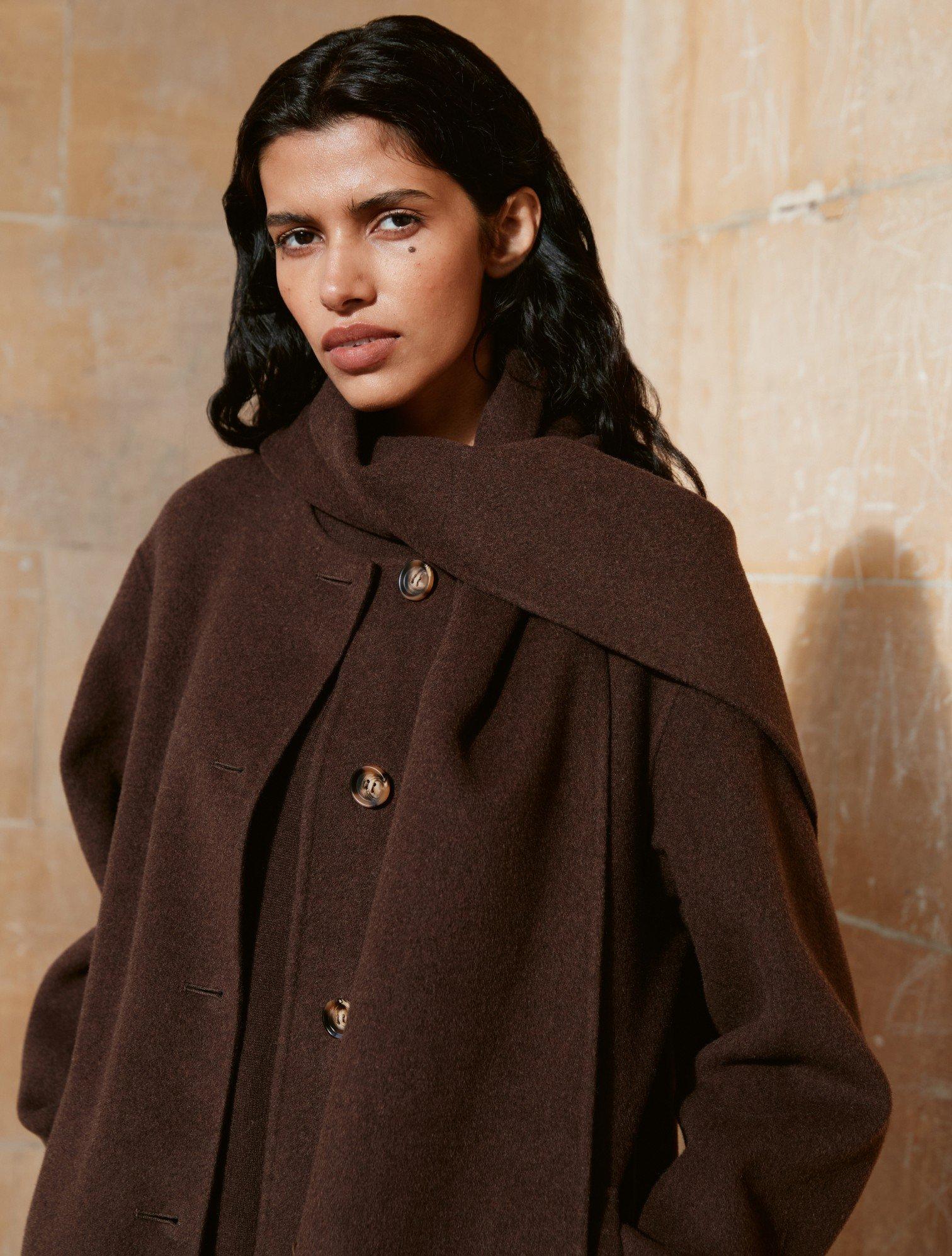 a woman in a brown coat standing in front of a wall