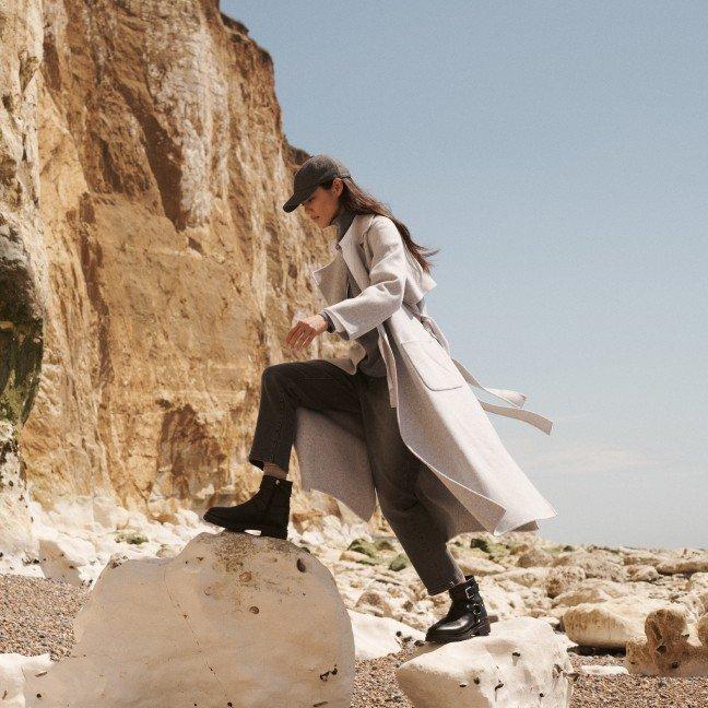 a woman in a trench coat and hat standing on a rock