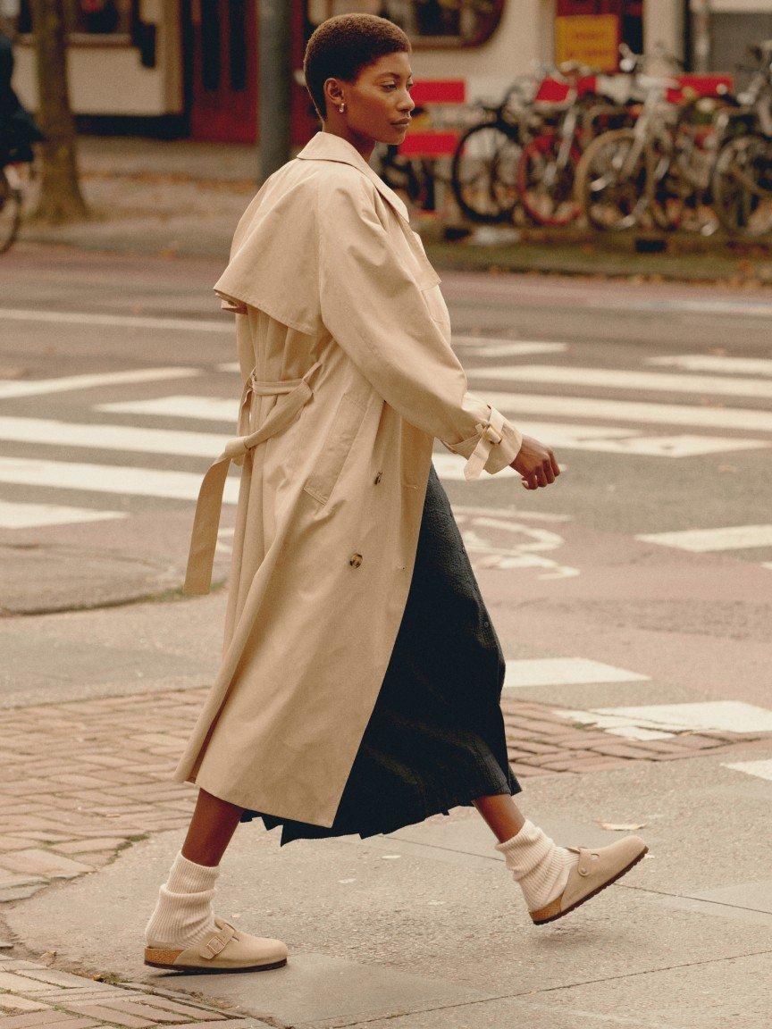 a woman in a trench coat crossing the street in front of a building
