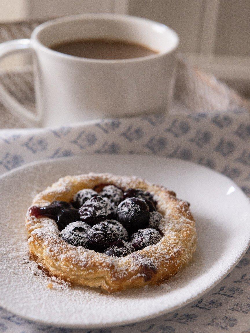 a pastry with blueberries on a white plate next to a cup of coffee