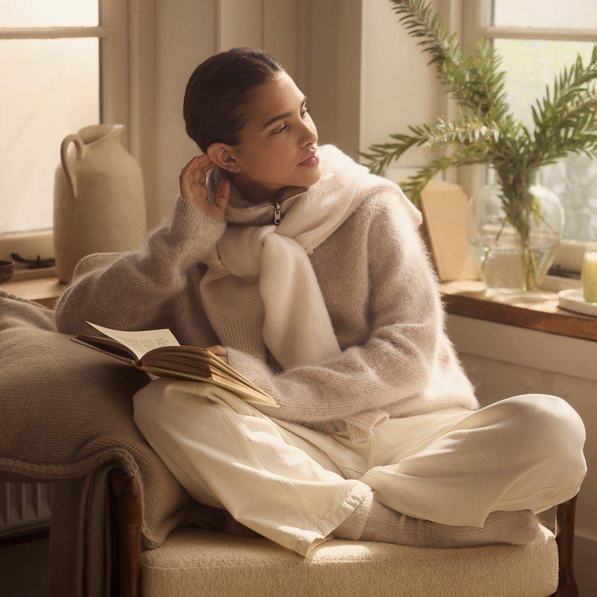 a woman in a sweater and scarf reading a book by a window