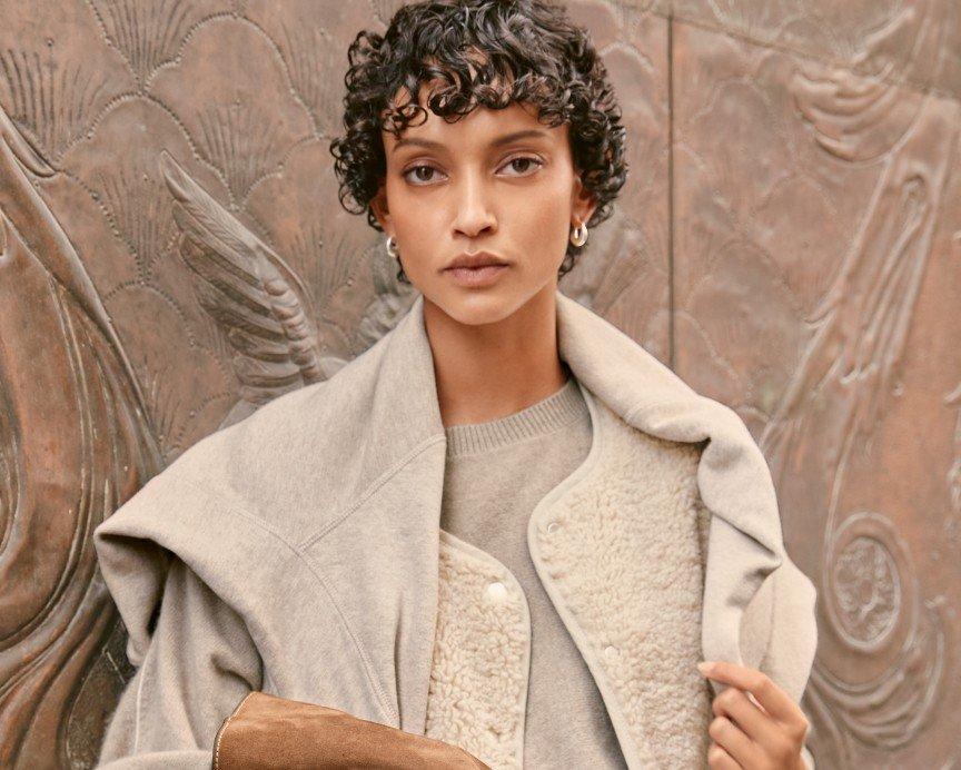 a woman in a white coat holding a purse in front of a wall