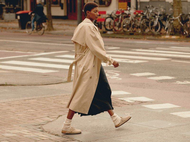 a woman walking across a street in a trench coat and sneakers