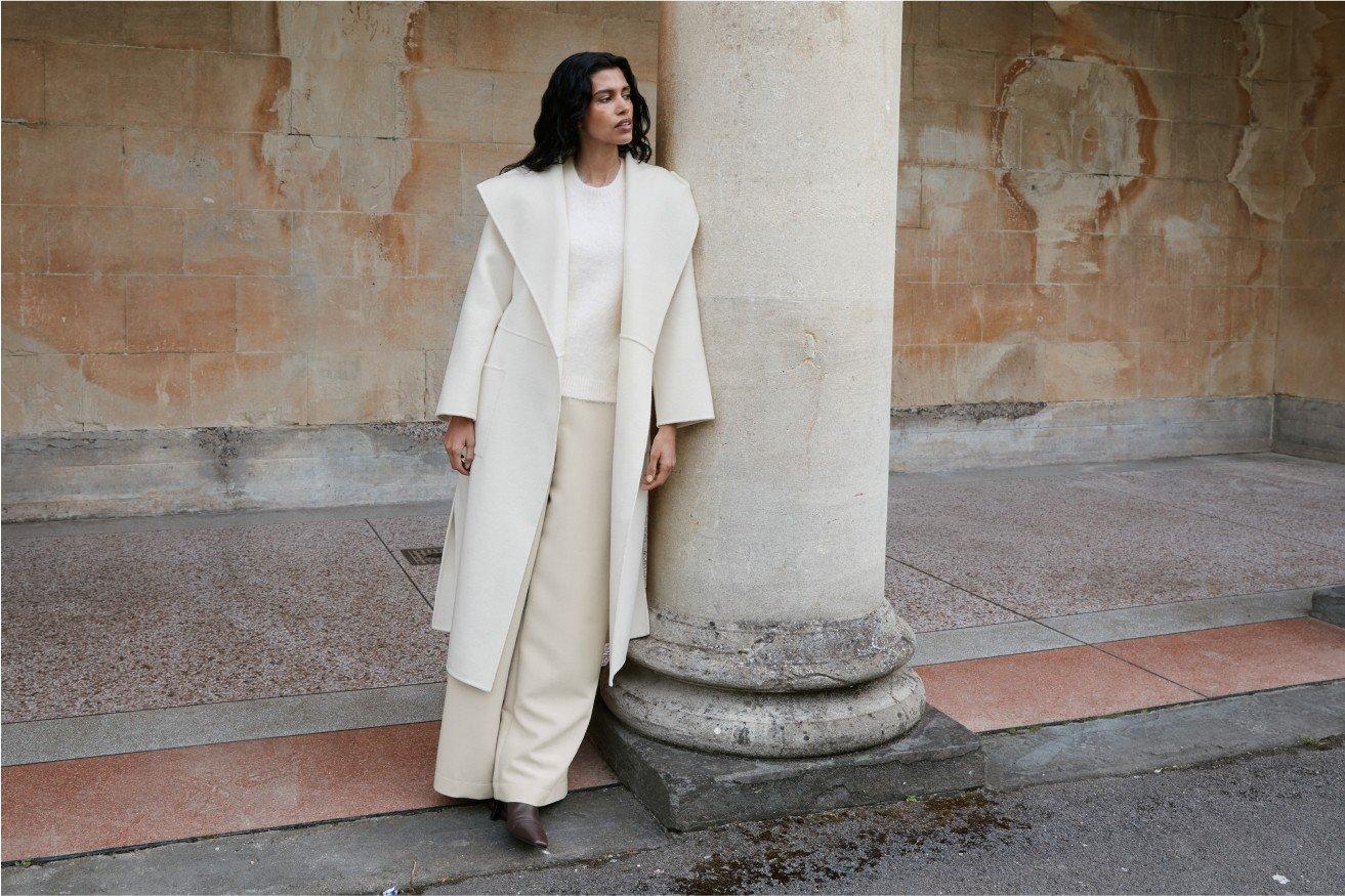 a woman in a white coat leaning against a pillar