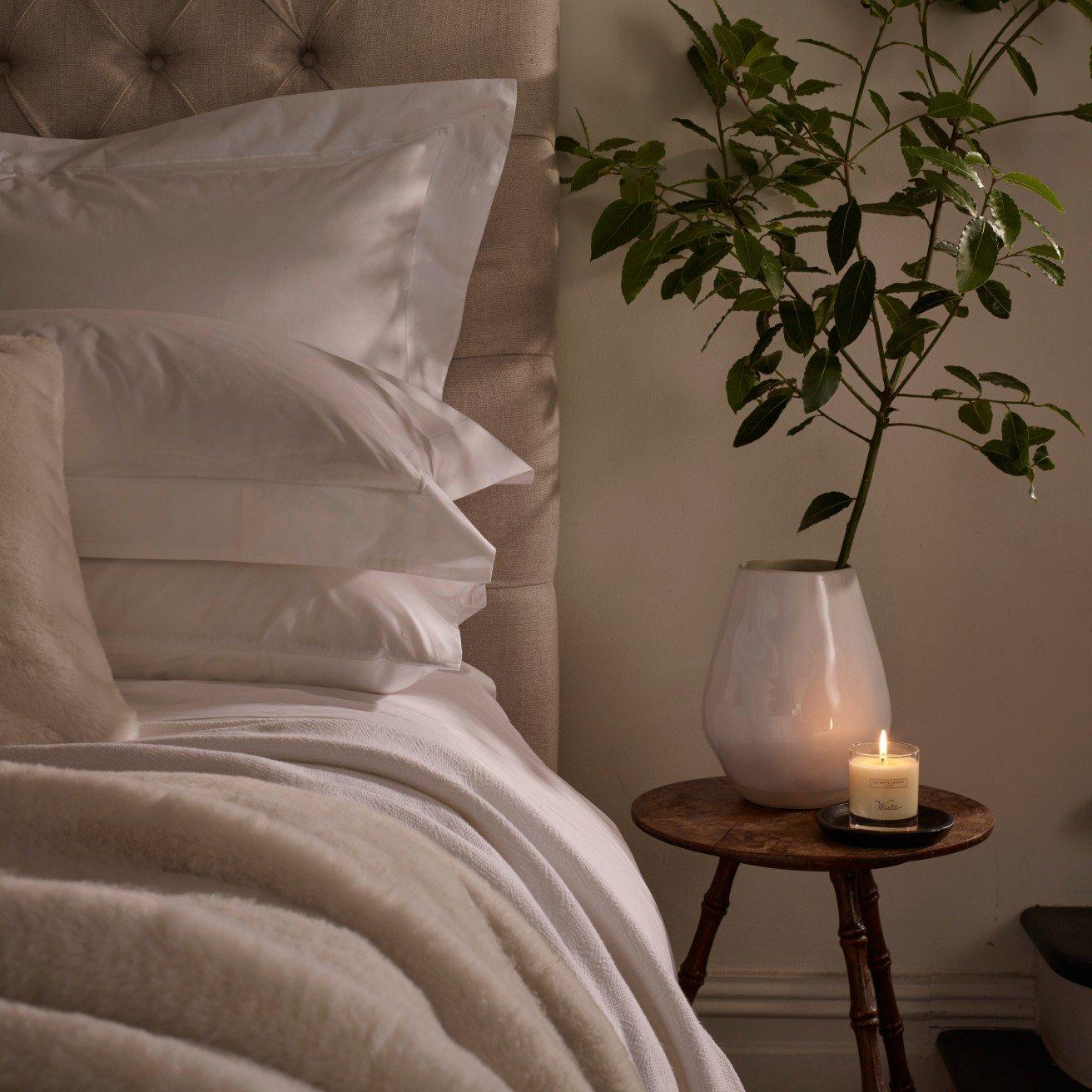 a candle on a small table next to a bed with a white comforter