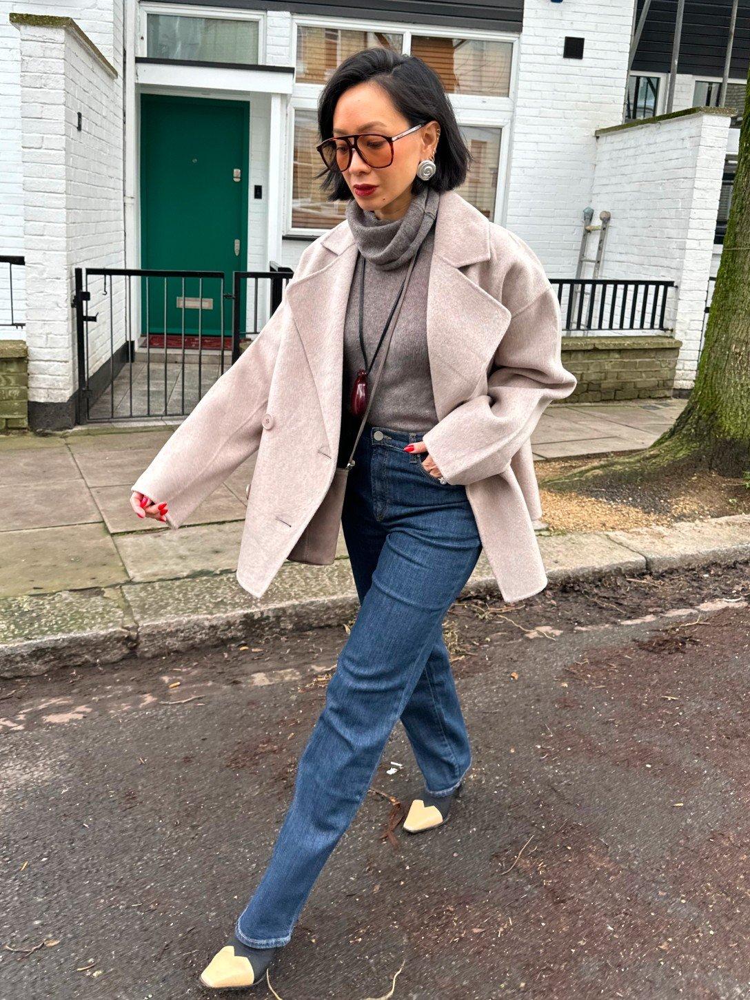 a woman in a beige coat and jeans walking down the street