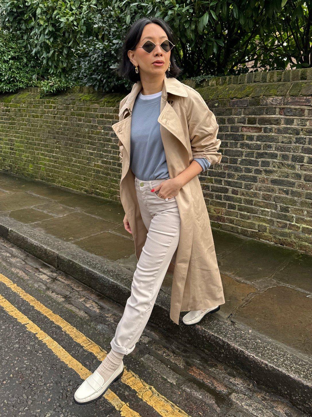 a woman in a trench coat and white pants is standing on the side of the road