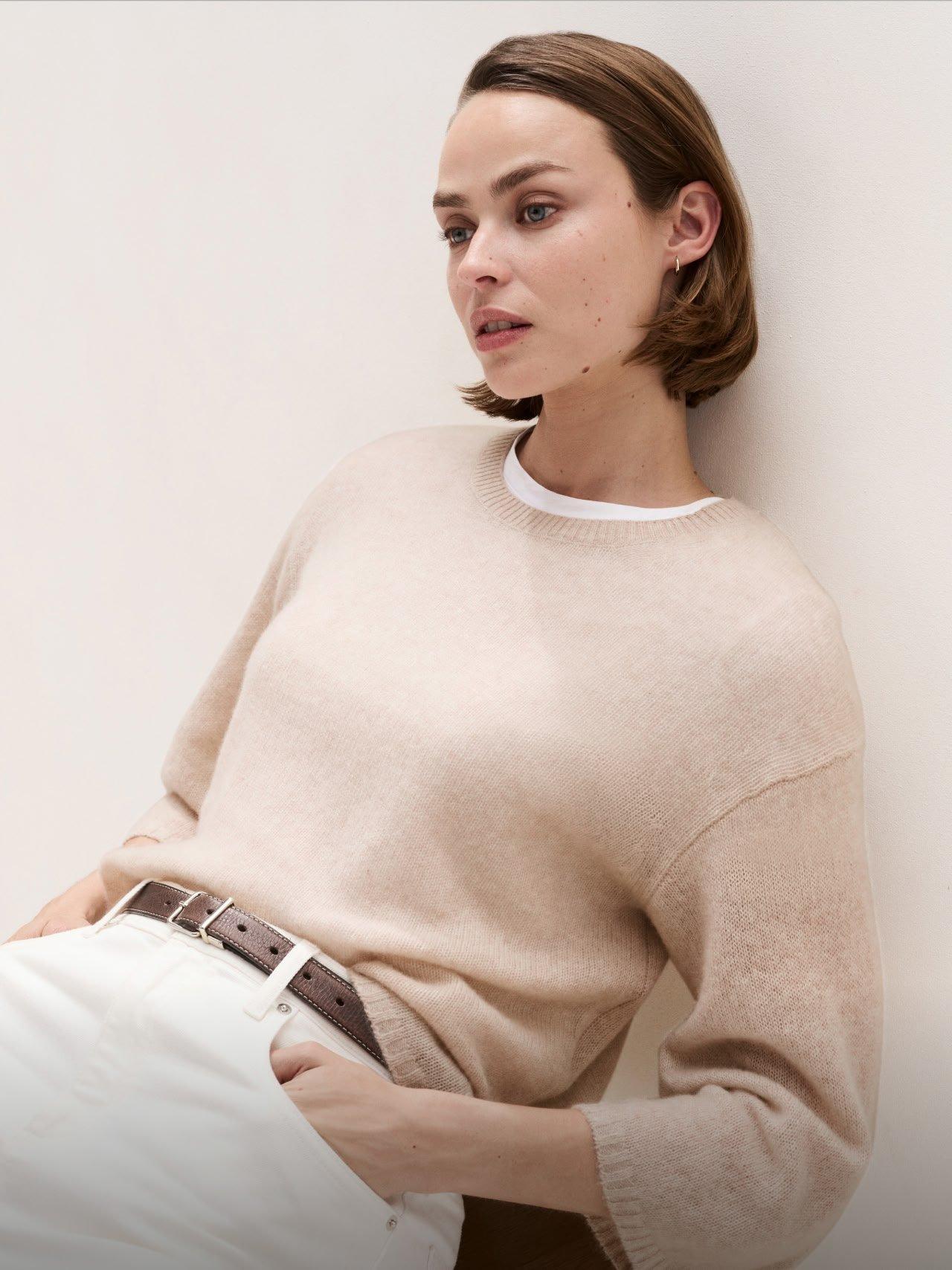 a woman leaning against a wall wearing a beige sweater