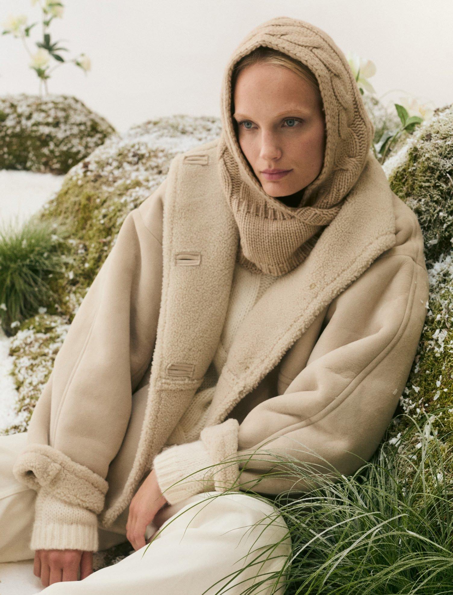 a woman in a beige coat sitting on a rock in the snow