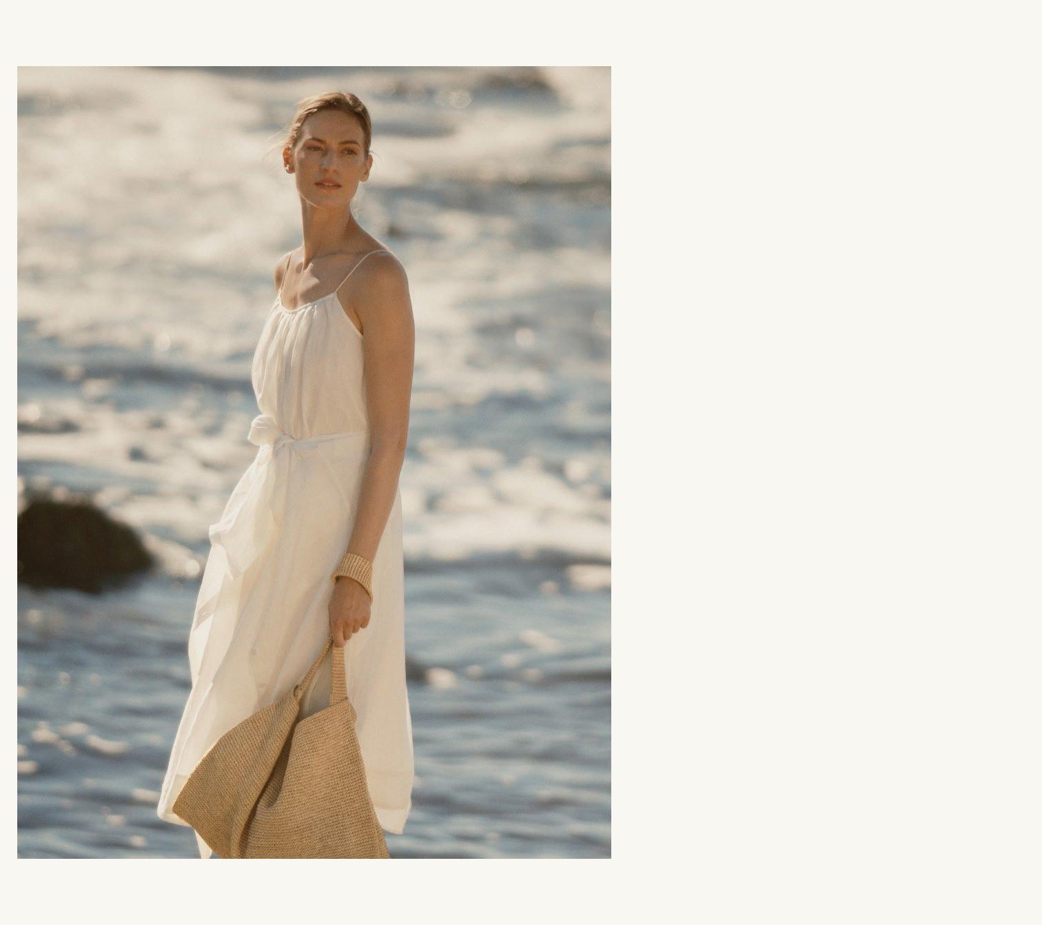 a woman in a white dress holding a straw bag by the water
