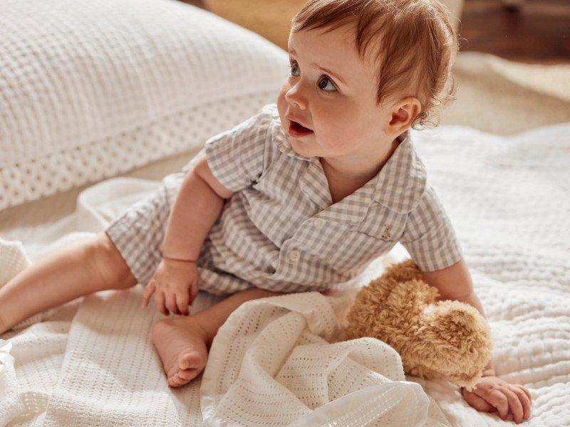 a baby in a white shirt sitting on a bed with a teddy bear