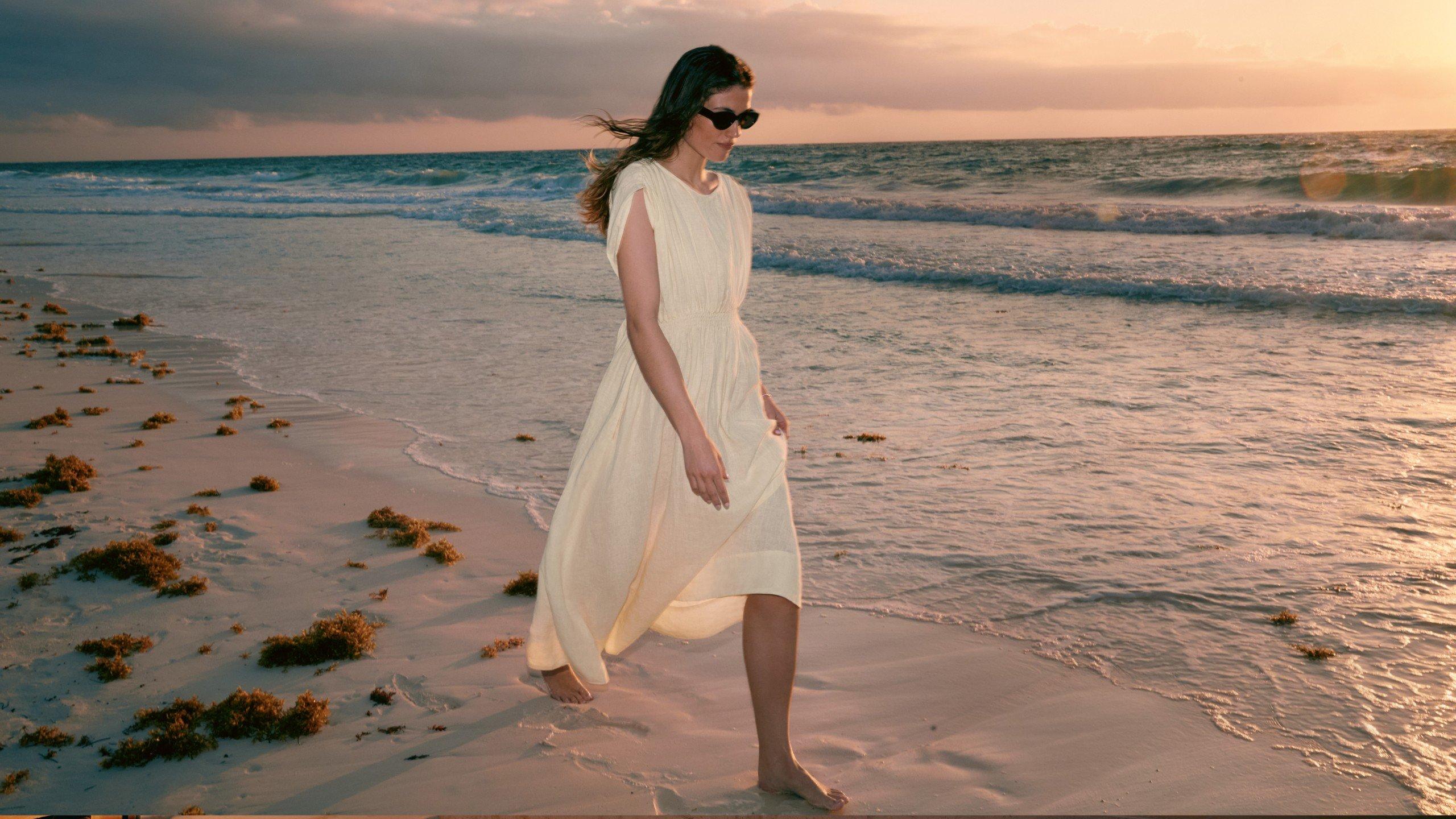 a woman in a white dress walking on the beach at sunset