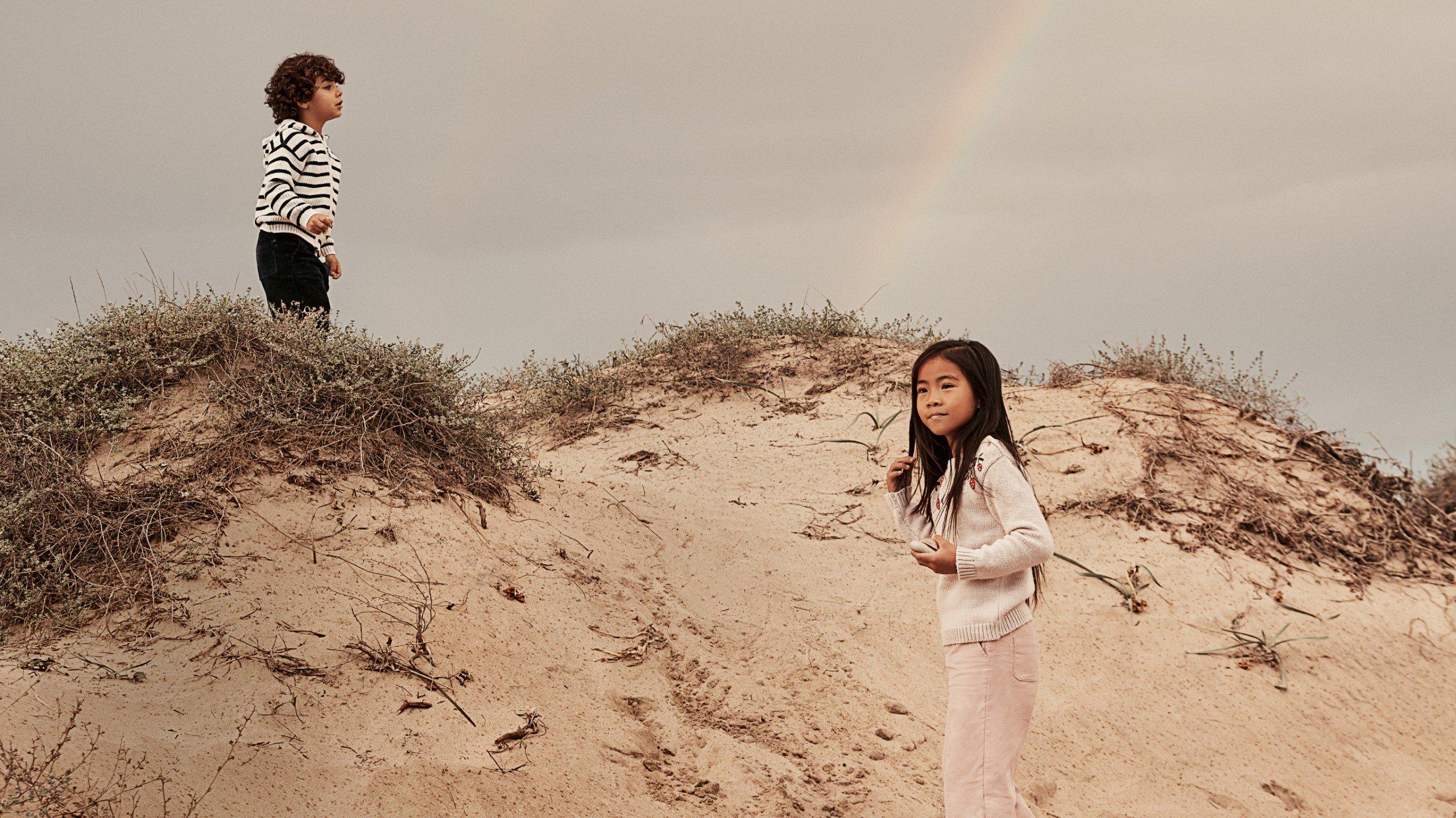 two children standing on a sandy hill with a rainbow in the sky