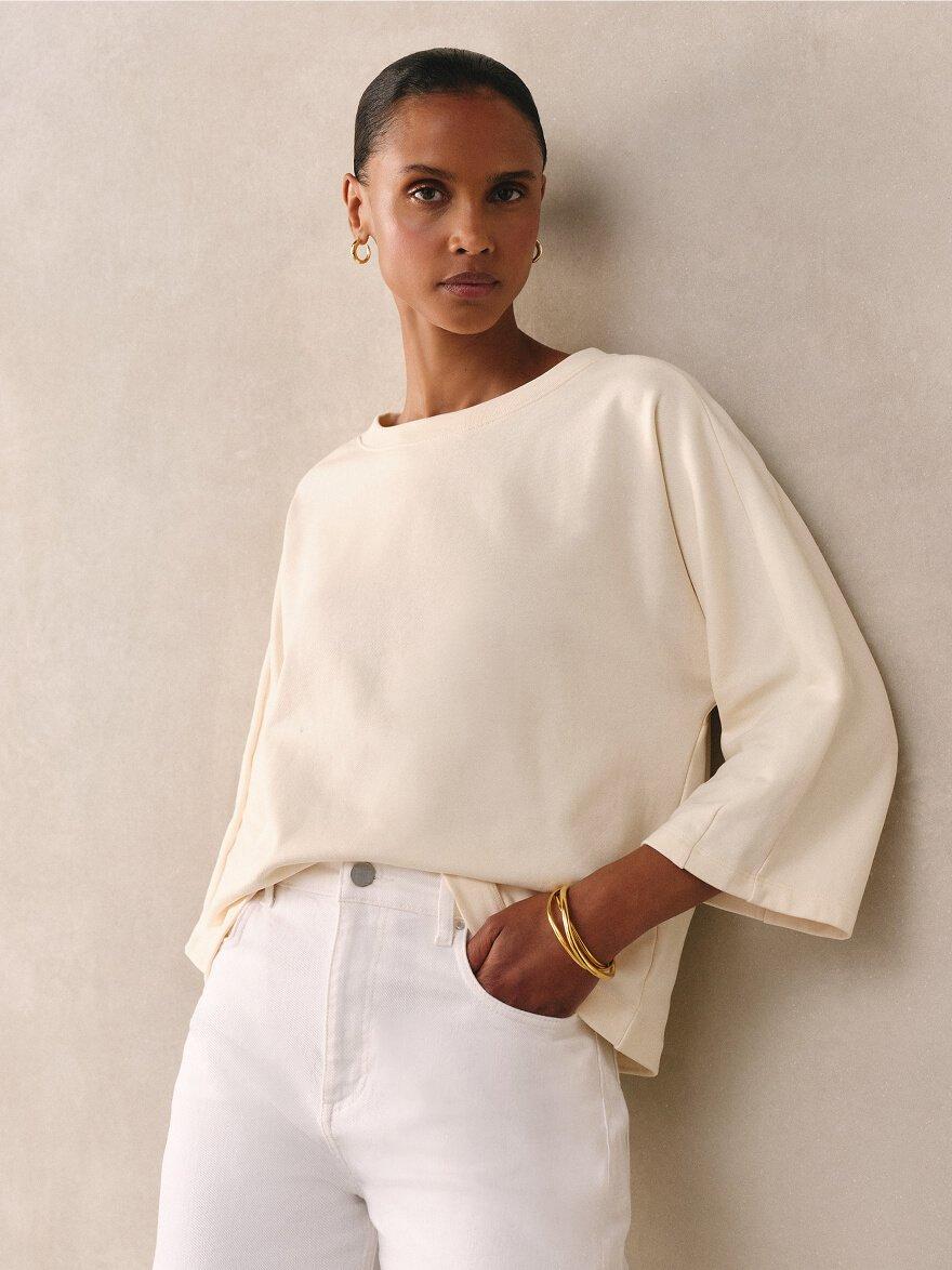 a woman in a tan sweater and white pants sitting on a stool