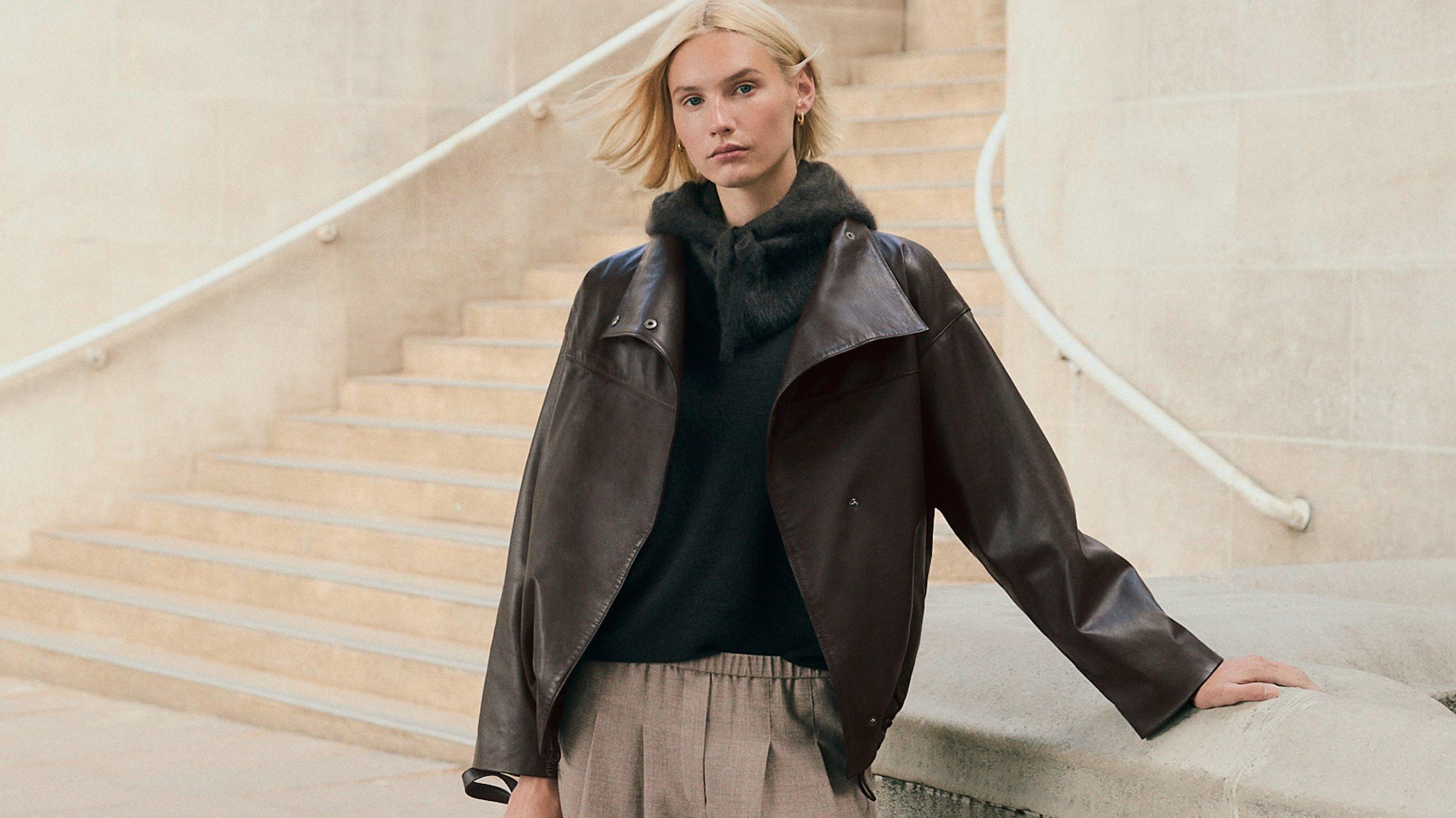 a woman in a black jacket and tan pants leaning against a wall