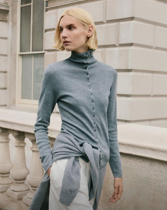 a woman in a grey sweater and white pants standing on a sidewalk